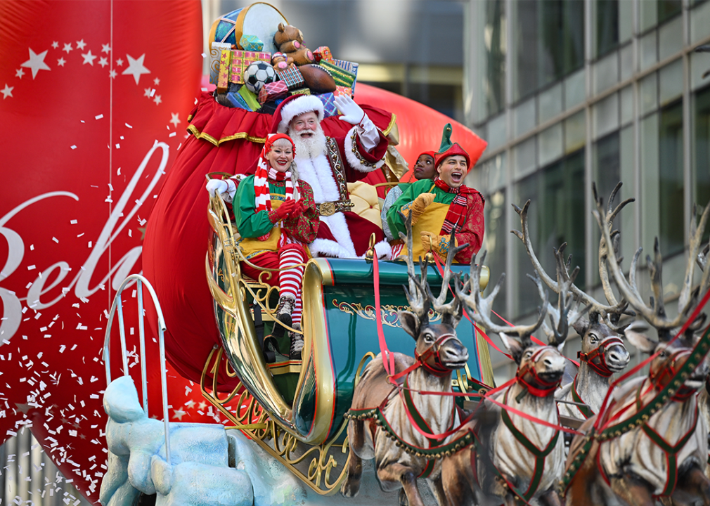 Santa Claus and an elf sit on a float in a sleigh pulled by reindeer.